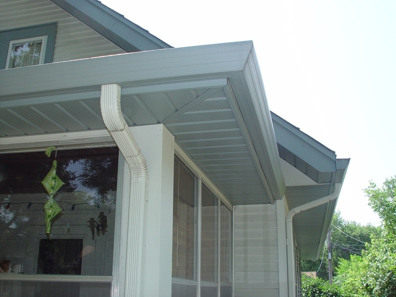 A close-up view of a house corner with white and gray siding showcases the precise work of a skilled contractor. The image highlights the roofline, gutters, and downspouts. A window is partially visible, adorned with green decorative ornaments inside, set against a backdrop of trees on a clear day.