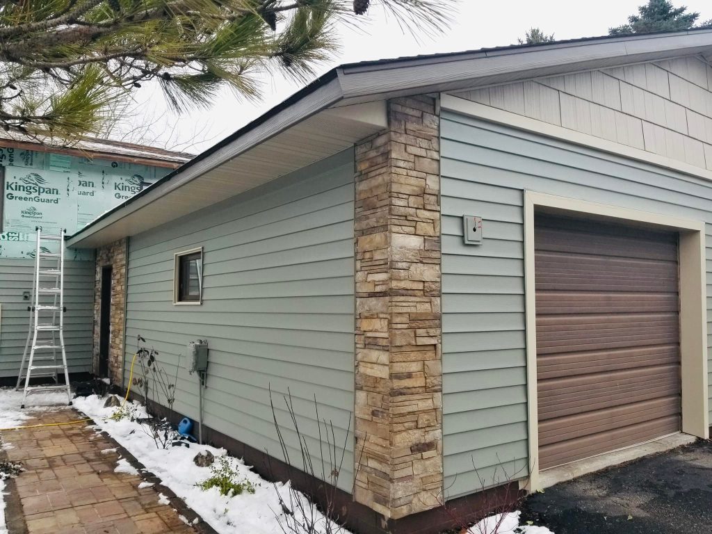 A newly constructed garage with light blue siding and stone accents showcases the work of an expert contractor. Snow blankets the ground, while a ladder leans against the building. The garage, featuring a brown door and abc seamless roofing, is set against a backdrop of trees.