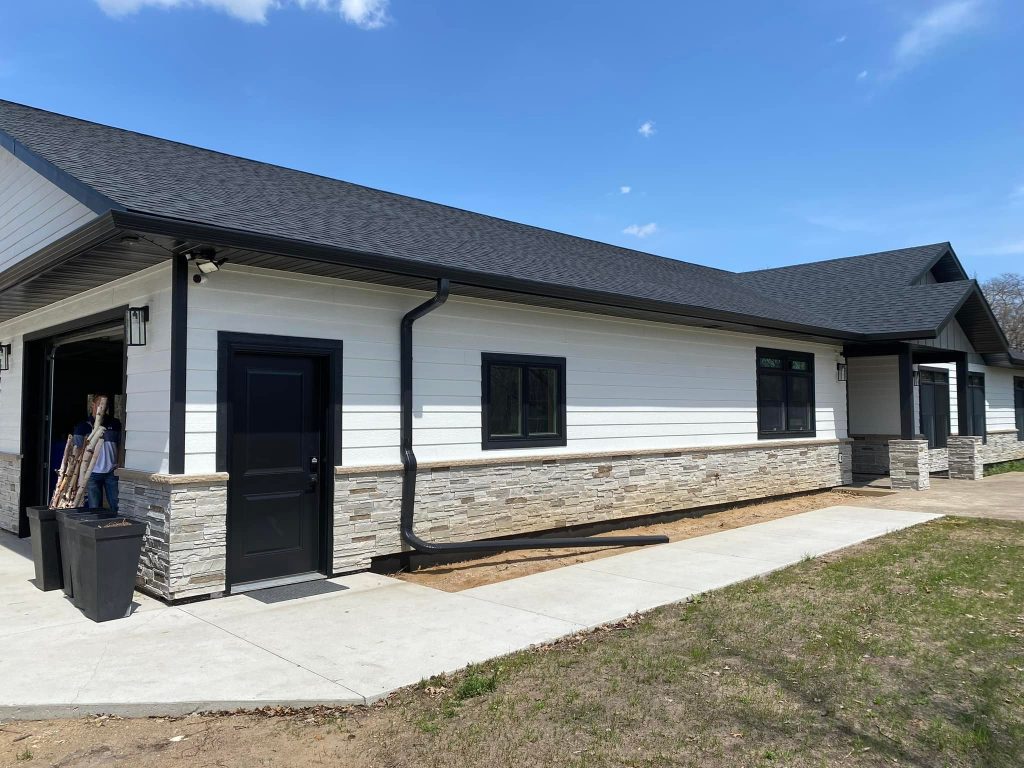 A modern single-story house features elegant white siding and stone accents, complemented by expert roofing in sleek black. The black trim around the windows and doors adds sophistication. A garage door is partially open, revealing a contractor inside. A driveway and lawn stretch invitingly in the foreground.