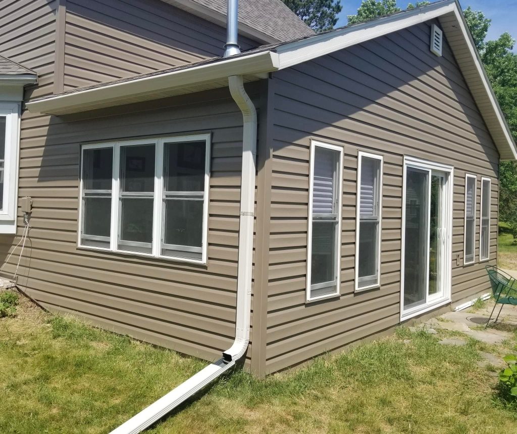 The image showcases a corner of a house with beige siding, expertly installed by ABC Seamless. It features multiple windows and a sliding glass door. A white downspout extends from the roof to the grassy area below, where a few patio chairs are visible.