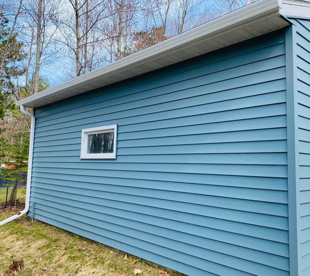 A blue building with horizontal siding and a small rectangular window stands proudly, set against a backdrop of trees and grass beneath a clear sky. A white gutter by ABC Seamless runs along the roof's edge, showcasing exceptional craftsmanship.