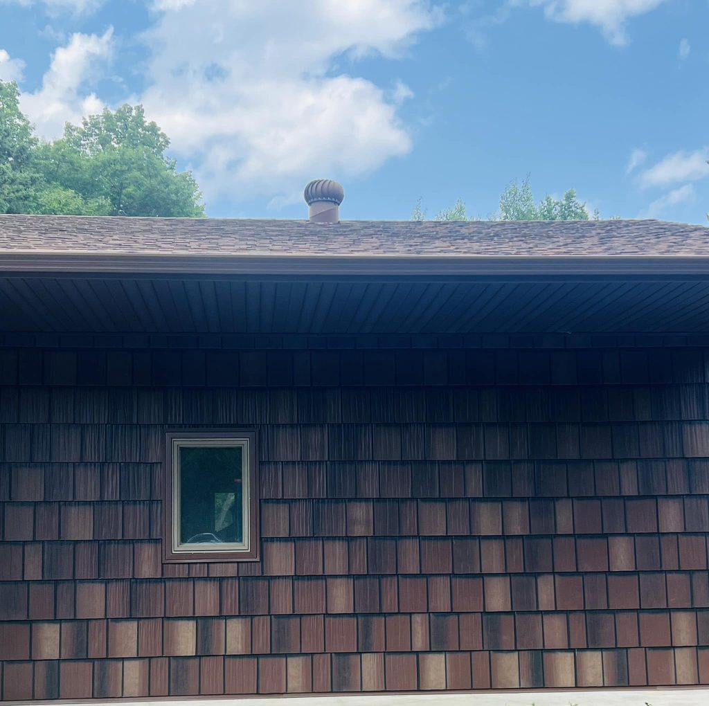 The image shows the side of a house with brown wooden shingles and a small window. A contractor-installed vent punctuates the roofing, while the sky above is partly cloudy with the tops of trees visible in the background.