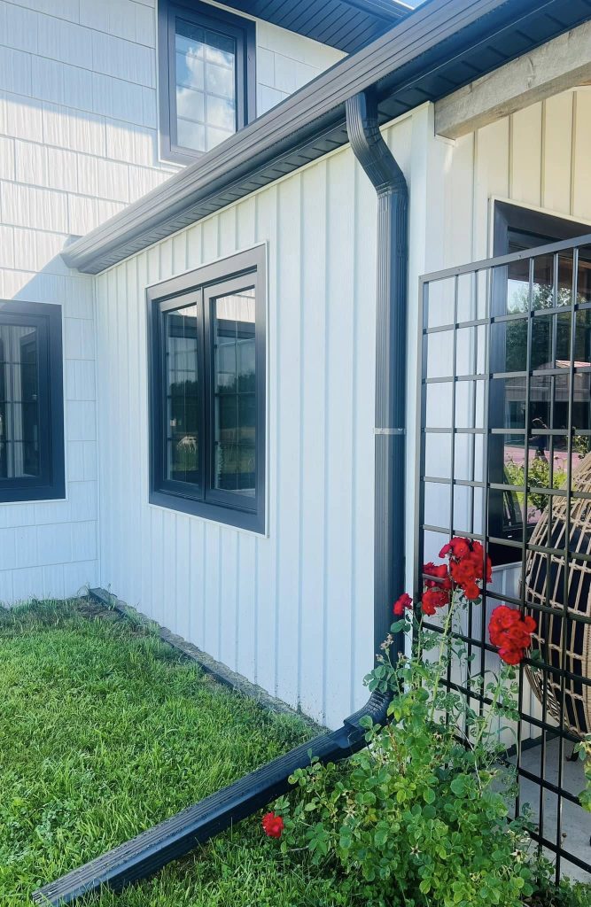 A white house with black window frames and sleek black guttering showcases the expert touch of a skilled contractor. Bright red flowers bloom near the corner, contrasting beautifully against the ABC Seamless siding. The yard is lush with green grass under a clear sky.