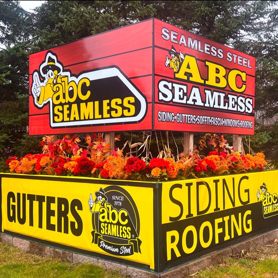 A colorful outdoor sign for "ABC Seamless" featuring bright red and yellow hues showcases their expert contractor services in seamless steel guttering, siding, and roofing. The sign is enveloped by lush greenery and vibrant autumn leaves.