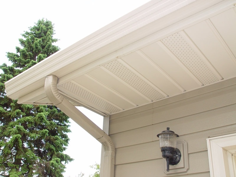 A close-up of a house's beige roof with soffit, fascia, and seamless siding by ABC Seamless. A downspout runs from the edge of the roof to the wall near vintage-style windows, accompanied by an exterior light fixture. A green pine tree is visible in the background.