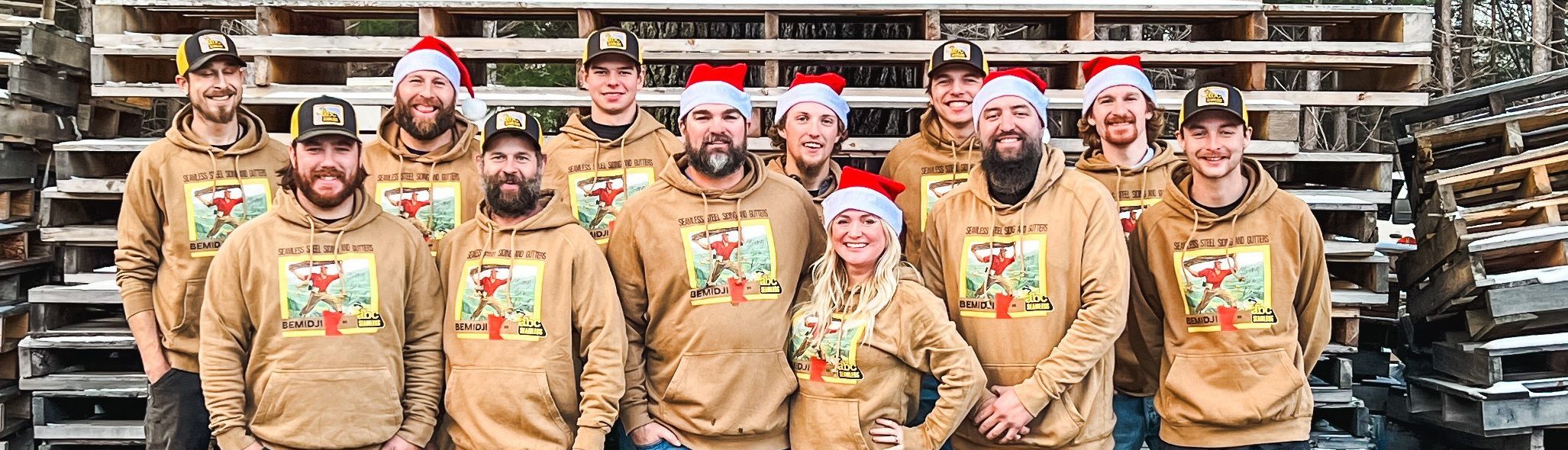 A group of twelve people wearing matching brown sweatshirts, some with black caps and others with red Santa hats, pose in front of stacked wooden pallets. They all smile, their shirts featuring a holiday-themed graphic that cleverly showcases their pride as a local roofing contractor team.