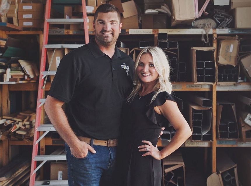 A man and a woman stand together smiling in a workshop, resembling seasoned contractors amidst shelves of materials. The man wears a black polo shirt and jeans, while the woman sports a sleek black dress. A red ladder is visible in the background, hinting at their expertise in windows and siding installations.