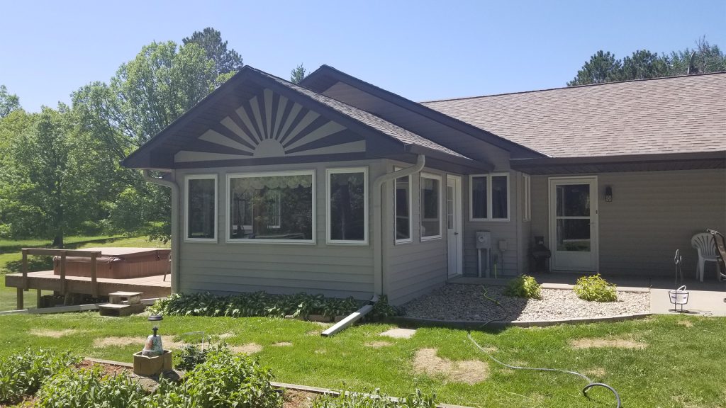 A single-story house with gray siding and expertly crafted roofing showcases a large sunburst design above three front windows. A wooden deck is on the left, and greenery surrounds the building under a clear, blue sky.