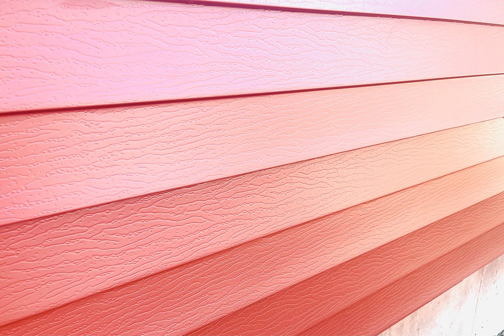 Close-up of textured, overlapping horizontal siding panels in various shades of pink and red, creating a gradient effect. The surface texture resembles a subtle wood grain pattern, perfectly complementing the ABC Seamless roofing above.