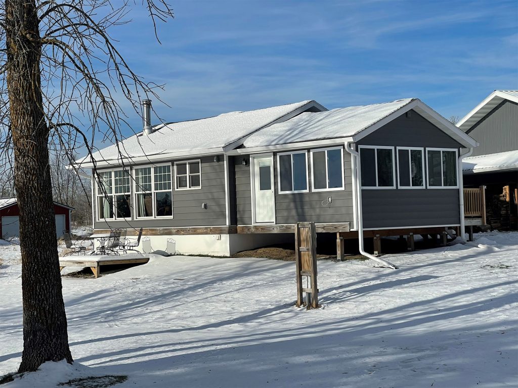 A gray house with white trim and large windows is surrounded by snow, showcasing its expertly crafted siding. The sky is clear and blue, highlighting a leafless tree standing in the foreground.