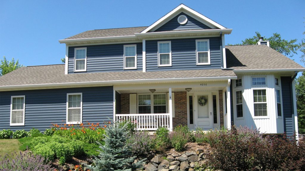 A two-story blue house with white trim and large windows, featuring a front porch and a well-maintained garden with various plants and shrubs. The clear blue sky complements the home's inviting appearance.