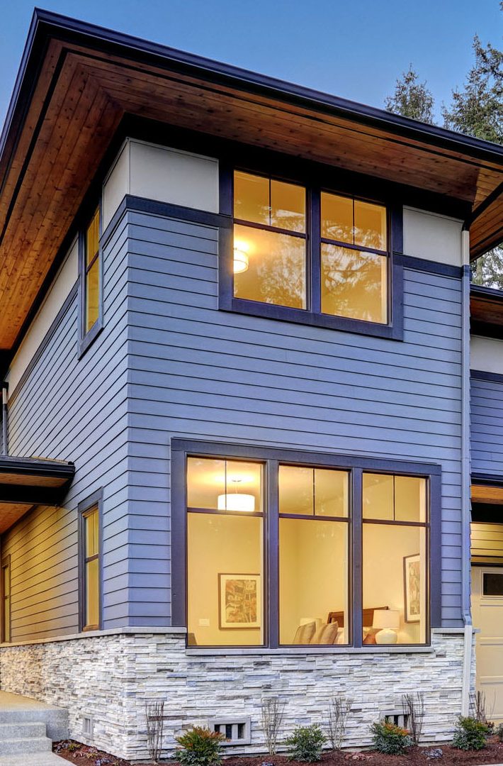 A modern house exterior boasts blue siding and large windows crafted by ABC Seamless. The stone-clad lower section complements the sleek roofing, while warm lights glowing through the windows add a cozy touch. A clear sky suggests it's early evening, enhancing the home's inviting ambiance.