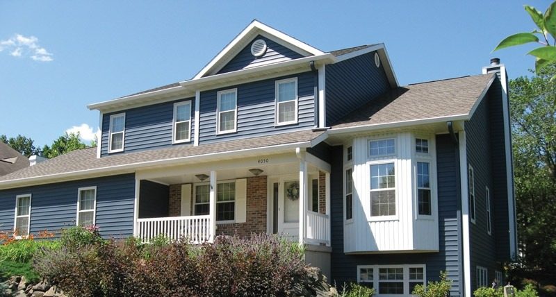 A charming two-story blue house in Bemidji, with white trim, features a covered porch and large bay window. The front yard boasts neatly landscaped grass, bushes, and orange flowers. ABC Seamless siding enhances its beauty, while trees and a clear blue sky complete the inviting scene.