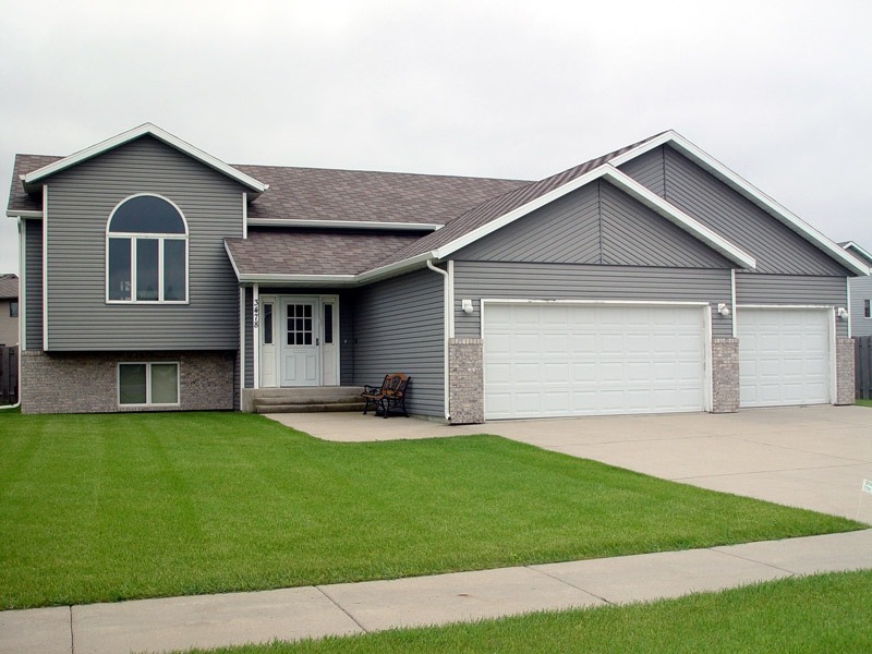 A modern suburban house with gray siding and a three-car garage. The front yard has a well-maintained green lawn. The house features a large arched window above the entrance and a small patio area with chairs near the front door.