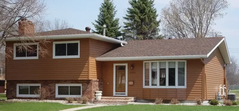 A split-level house with brown siding and a brick foundation. It has a large front window and a chimney. The yard has a few small shrubs, and there are tall trees in the background under a clear sky.