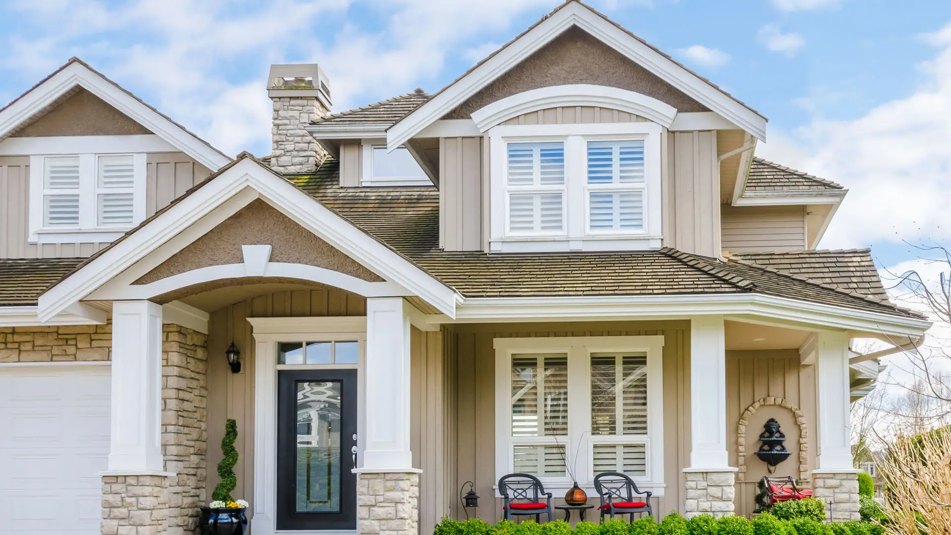 A charming two-story house with beige siding and stone accents features a covered porch, black front door, and white window shutters. Energy-efficient windows enhance the home's appeal while improving comfort. Two red chairs amid green shrubbery invite relaxation under a partly cloudy sky.