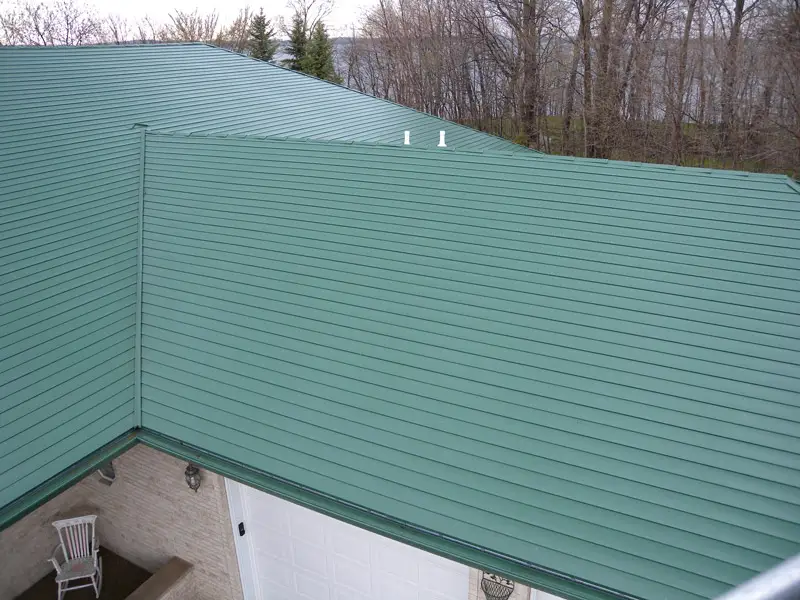 A house with a green metal roof viewed from above. The roof has a smooth, ridged texture and covers a white garage door. Bare trees are visible in the background, indicating a winter or early spring setting. A white chair is seen on a porch below.