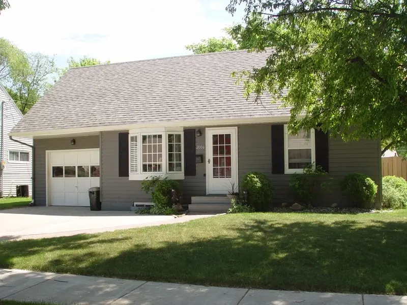 A small, gray one-story house with steel siding and a sloped roof offers robust home protection against harsh weather. Featuring white trim, black shutters, a single garage, and a front porch with a white door adorned by green shrubs. A manicured lawn leads to the sidewalk, framed by trees beyond.